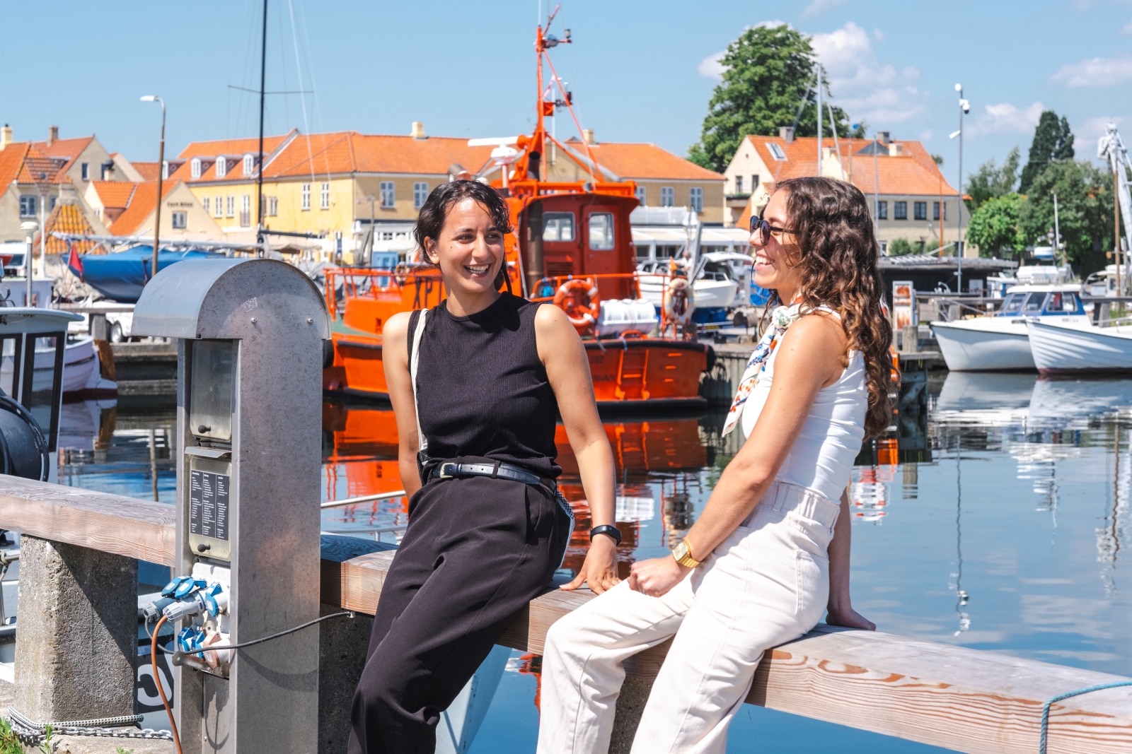 To kvinder sidder på rækværk på havnen